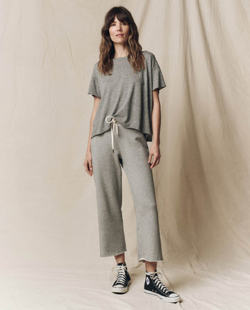 A person in THE GREAT. CORE KNITS' Wide Leg Cropped Sweatpant in Varsity Grey stands against a beige backdrop wearing black high-top sneakers.