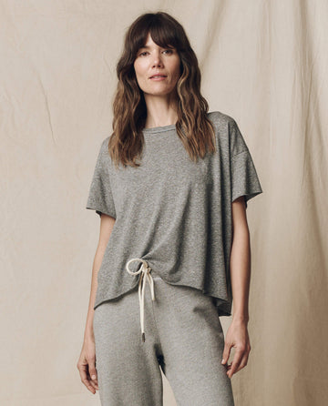 A person in THE GREAT. CORE KNITS' Heather Grey Crop Tee and matching drawstring pants stands against a beige fabric backdrop.