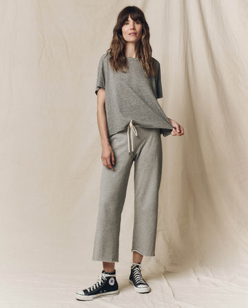 A woman in The Crop Tee - Heather Grey by THE GREAT. CORE KNITS poses against a beige backdrop, casually holding the hem of her super soft cotton top while wearing black sneakers.