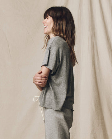 A person with long brown hair smiles in The Crop Tee, Solid--Heather Grey by THE GREAT. CORE KNITS, standing sideways against a beige fabric backdrop.