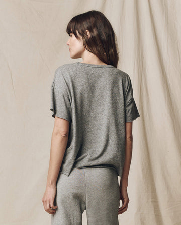 A person with long hair stands sideways, wearing The Crop Tee in Heather Grey by THE GREAT. CORE KNITS, made of super soft cotton, along with pants. The background is plain beige fabric.