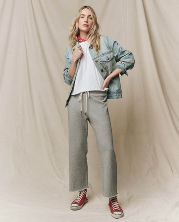 Person wearing a denim jacket, The Crop Tee in True White by THE GREAT. CORE KNITS, gray sweatpants, and red sneakers, standing against a beige backdrop.