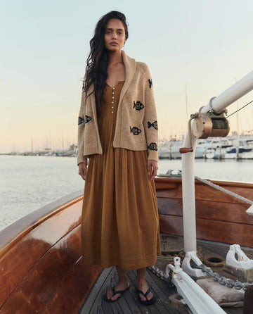 A woman stands on a wooden boat wearing THE GREAT. SU26 The Day Break Dress in Clay, paired with a beige fish-motif cardigan and black sandals; marina and docked boats appear in the background.
