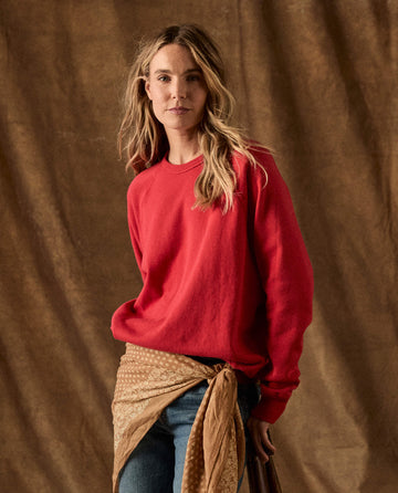 A woman wears a long-sleeved red The Fleece College Sweatshirt -- Campfire with blue jeans and a patterned brown scarf tied at her waist.