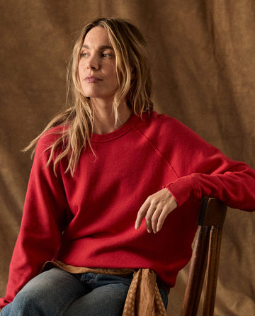 A person wearing a red raglan-sleeve The Fleece College Sweatshirt in Campfire, sitting on a wooden chair.