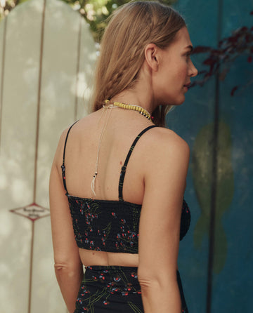 A woman with long blonde hair stands outdoors, facing away from the camera, wearing THE GREAT. SP26 SWIM's The Reversible Cinched Bandeau in Windblown Bouquet and a yellow beaded necklace.