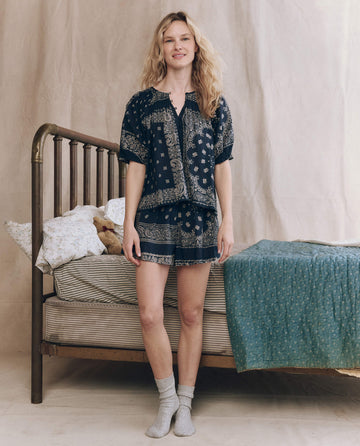 A woman stands in front of a metal bed wearing THE GREAT. F25 SLEEP's The Smocked Sleep Short in Bandana Patchwork and gray socks. The bed features patterned bedding, with a teddy bear beside the pillow.