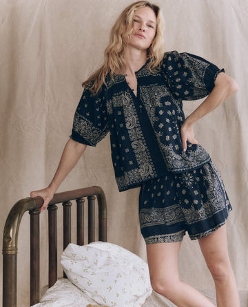 A woman in THE GREAT. F25 SLEEP The Smocked Sleep Top – Bandana Patchwork stands beside a brass bed with white floral bedding, set against a neutral fabric backdrop.