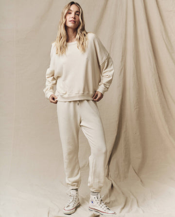 A person with long blonde hair is standing in front of a beige draped background, wearing The Teammate Sweatshirt in Washed White from THE GREAT. FALL21 KNITS, matching sweatpants, and white high-top sneakers.
