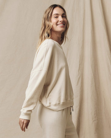 Person standing and smiling at the camera, wearing The Teammate Sweatshirt in Washed White, featuring a vintage look and a relaxed fit, against a beige backdrop.