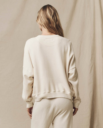 A person with long hair is standing and facing away from the camera, wearing a relaxed fit sweatshirt named The Teammate Sweatshirt in Washed White from THE GREAT. FALL21 KNITS, paired with matching pants against a plain, beige background.