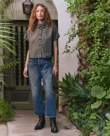 Outdoors by greenery, someone with wavy hair wears THE GREAT. PF25 The Valor Top — Smoke Wash short-sleeve button-up shirt, blue jeans, and black boots. Casual style ideal for everyday wear keywords.