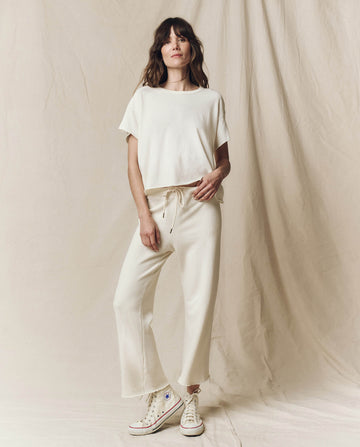 A person stands against a beige backdrop, wearing The Crop Tee in washed white by THE GREAT. CORE KNITS and high-rise jeans, paired with white high-top sneakers.