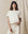 Wearing The Crop Tee in Washed White by THE GREAT. CORE KNITS and high-rise jeans, a person stands against a beige background, exuding effortless style.