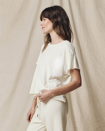 A person wearing "The Crop Tee. Solid -- Washed White" by THE GREAT. CORE KNITS stands against a draped fabric background, looking to the side.