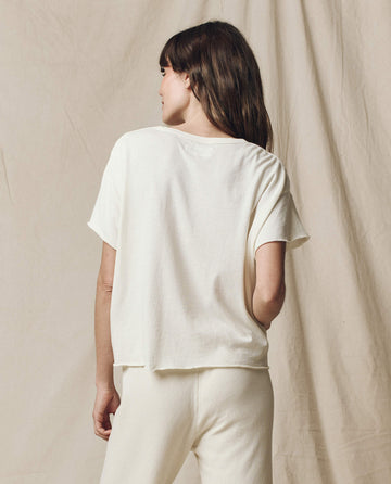 A person with long brown hair stands in front of a beige fabric backdrop, wearing THE GREAT. CORE KNITS' The Crop Tee in Washed White and super soft cotton pants.