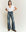 A person in a casual outfit stands against a plain background, wearing a white V-neck T-shirt, The Dock Jean in Marina Wash by THE GREAT. PF24, and brown shoes.