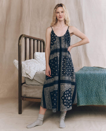 A woman in THE GREAT. F25 SLEEP The Slip Sleep Dress in Bandana Patchwork and gray socks stands by a vintage brass bed with patterned bedding in a neutral-toned room.