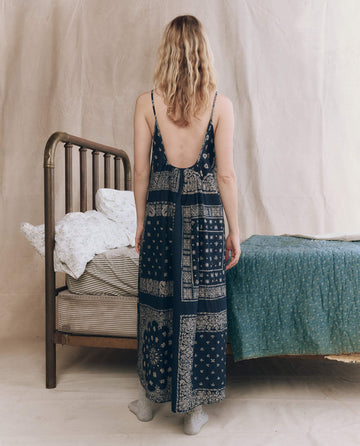 A woman with long blond hair stands with her back to the camera, wearing THE GREAT. F25 SLEEP's The Slip Sleep Dress in Bandana Patchwork beside a bed with patterned bedding in a softly lit room.