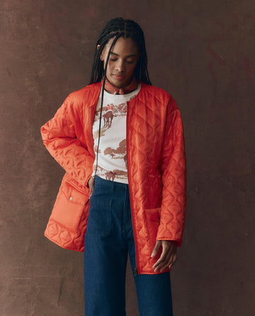 A person with long braids wears THE GREAT. F25 D1 The Quilted Liner Parka in Army with Hot Orange over a printed white t-shirt and blue jeans, standing against a brown backdrop.