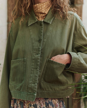 A person wearing THE GREAT. PF25 The Rancher Jacket - Classic Army, styled with a beige patterned turtleneck and floral skirt.