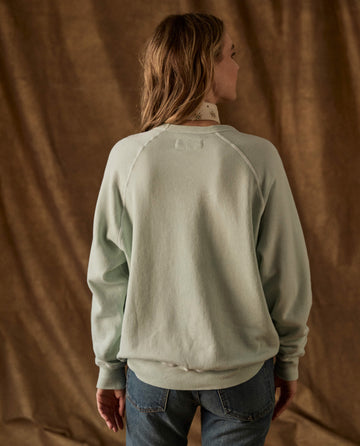 Back view of a THE GREAT. H26 FLEECE BACK KNITS The Fleece College Sweatshirt in Fresh Mint, featuring raglan sleeves and worn over a patterned collared shirt and blue jeans.