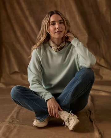 A person wearing The Fleece College Sweatshirt in Fresh Mint and blue jeans sitting cross-legged.