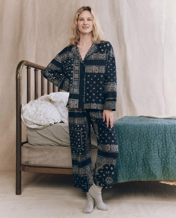 A person stands by a bed in THE GREAT. F25 SLEEP The Easy Sleep Pant – Bandana Patchwork and gray socks, with one hand resting on the bed frame.