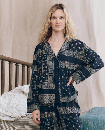 A woman with wavy blonde hair stands by a bed wearing THE GREAT. F25 SLEEP’s The Pajama Shirt. -- Bandana Patchwork, paired with matching pants for a vintage-inspired sleepwear look.