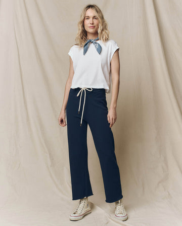 A woman with long hair stands against a beige draped backdrop wearing THE GREAT. FALL 23 KNITS' The Wide Leg Cropped Sweatpant in True Navy, paired with a white top. Her sneakers and a bandana tied around her neck enhance the casual vibe.