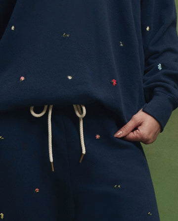 A person models THE GREAT. PS26’s The Stadium Sweatpant. Embroidered—Navy with Tossed Floral Embroidery—featuring small colorful florals and beige drawstrings, evoking vintage athletic style against a green backdrop.
