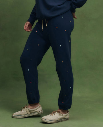Model wears THE GREAT. PS26 The Stadium Sweatpant in navy with tossed floral embroidery, paired with beige sneakers, standing before a green background. Inspired by vintage athletic wear and detailed with delicate embroidered florals.
