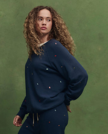 A person with curly hair wears THE GREAT. PS26 The College Sweatshirt in navy with tossed floral embroidery, paired with matching pants, standing before a green background and looking to the side.
