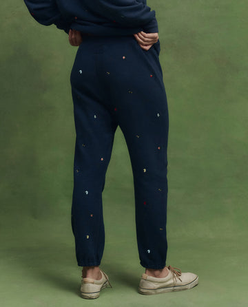 A person stands on a green background wearing THE GREAT. PS26 The Stadium Sweatpant. Embroidered—navy with tossed floral embroidery—paired with a matching sweatshirt and beige lace-up shoes; classic vintage athletic wear made in Los Angeles.