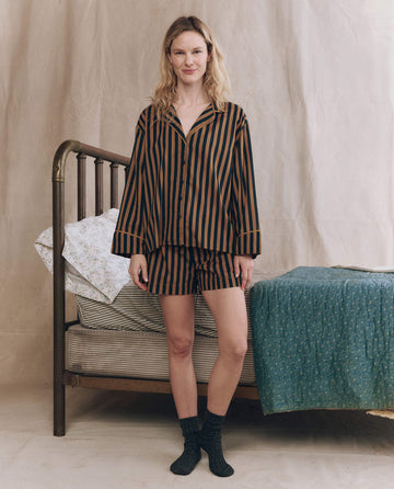 A woman stands indoors by a bed, wearing THE GREAT. F25 SLEEP "The Pajama Shirt" in Club Stripe—black and brown striped cotton sleepwear with shorts and socks. The bed features striped sheets, a floral pillow, and a blue blanket.