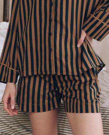 Model wearing The Sleep Boxer Short in Club Stripe by THE GREAT. F25 SLEEP, featuring brown and black vertical stripes with an elasticated waistband—one hand on hip, the other at their side.