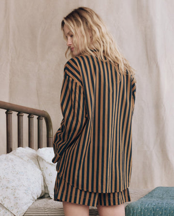 A woman with long blonde hair stands by a bed wearing THE GREAT. F25 SLEEP's The Sleep Boxer Short in Club Stripe—black and brown vertical striped cotton boxer shorts with an elastic waist—facing slightly away from the camera.