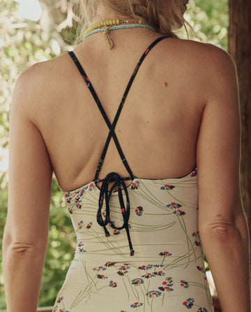 A woman stands with her back to the camera, wearing THE GREAT. SP26 SWIM's The Reversible Keyhole One Piece in Windblown Bouquet with thin crisscross straps and a floral pattern.