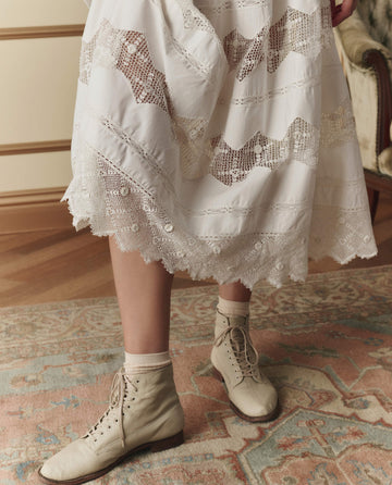 A person wears THE GREAT. SP26 D2 The Rosette Crochet Canary Skirt in cream, paired with cream socks and beige lace-up ankle boots, standing on a patterned rug over wood flooring.