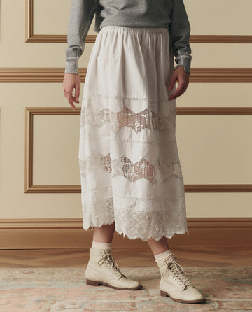 A person stands on a patterned rug wearing a gray long-sleeve top, cream socks, beige lace-up ankle boots, and THE GREAT. SP26 D2 The Rosette Crochet Canary Skirt in Cream.