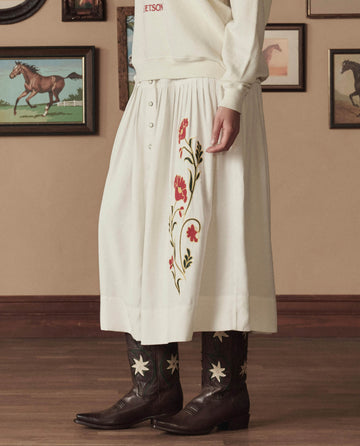 A person stands indoors wearing THE GREAT. STETSON SP26 The Cowboy Boot (Leather Floral) in Brown Floral and a white embroidered skirt, with framed horse pictures on the wall behind them.