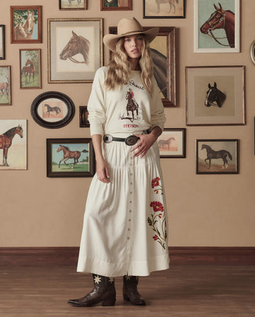 A woman wears THE GREAT. STETSON SP26's College Sweatshirt With Stetson Graphic in Washed White, paired with an embroidered skirt and cowboy boots, standing before a wall of framed horse pictures.