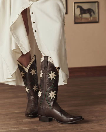 A person in a white dress wears THE GREAT. STETSON SP26 The Cowboy Boot (Leather Floral) in Brown Floral, standing on a wooden floor with framed pictures visible on the wall behind them.