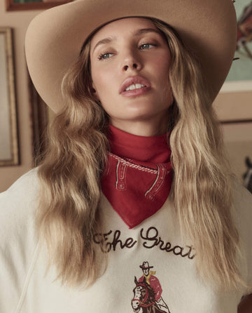 A person with long wavy blonde hair wears THE GREAT. STETSON SP26’s Stetson Western Bandana in red and a cream sweater featuring decorative text and an illustrated cowboy.