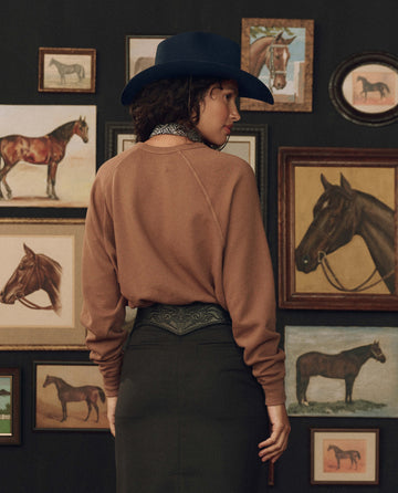 Wearing THE GREAT. F26 STETSON’s The College Sweatshirt w/ Stetson Graphic in Tobacco, a person stands facing a wall lined with framed horse paintings and illustrations, paired with a dark skirt and blue hat.