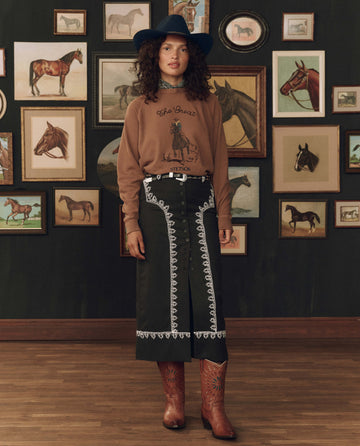 Woman in a cowboy hat, The College Sweatshirt w/ Stetson Graphic in Tobacco by THE GREAT. F26 STETSON, black skirt with white embroidery, and brown boots stands in front of a wall decorated with horse paintings.