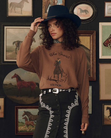A woman in a cowboy hat and western-style outfit stands in front of a wall adorned with framed horse paintings, wearing THE GREAT. F26 STETSON’s The College Sweatshirt w/ Stetson Graphic in Tobacco.