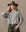 A person with long, wavy blonde hair wears THE GREAT. H25 COLLECTION's The Crochet Collar Pullover in Heather Grey, paired with a brown wide-brimmed hat and green belted pants, standing against a neutral background.