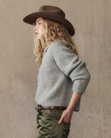 A person with long curly hair wears THE GREAT. H25 COLLECTION’s The Crochet Collar Pullover in Heather Grey, a brown wide-brim hat, and green pants, standing in profile against a neutral background.