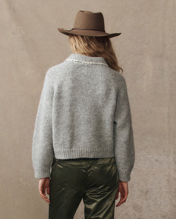 Person with long hair wears THE GREAT. H25 COLLECTION’s The Crochet Collar Pullover in Heather Grey, paired with a brown hat and olive green pants, standing back to the camera against a neutral background.